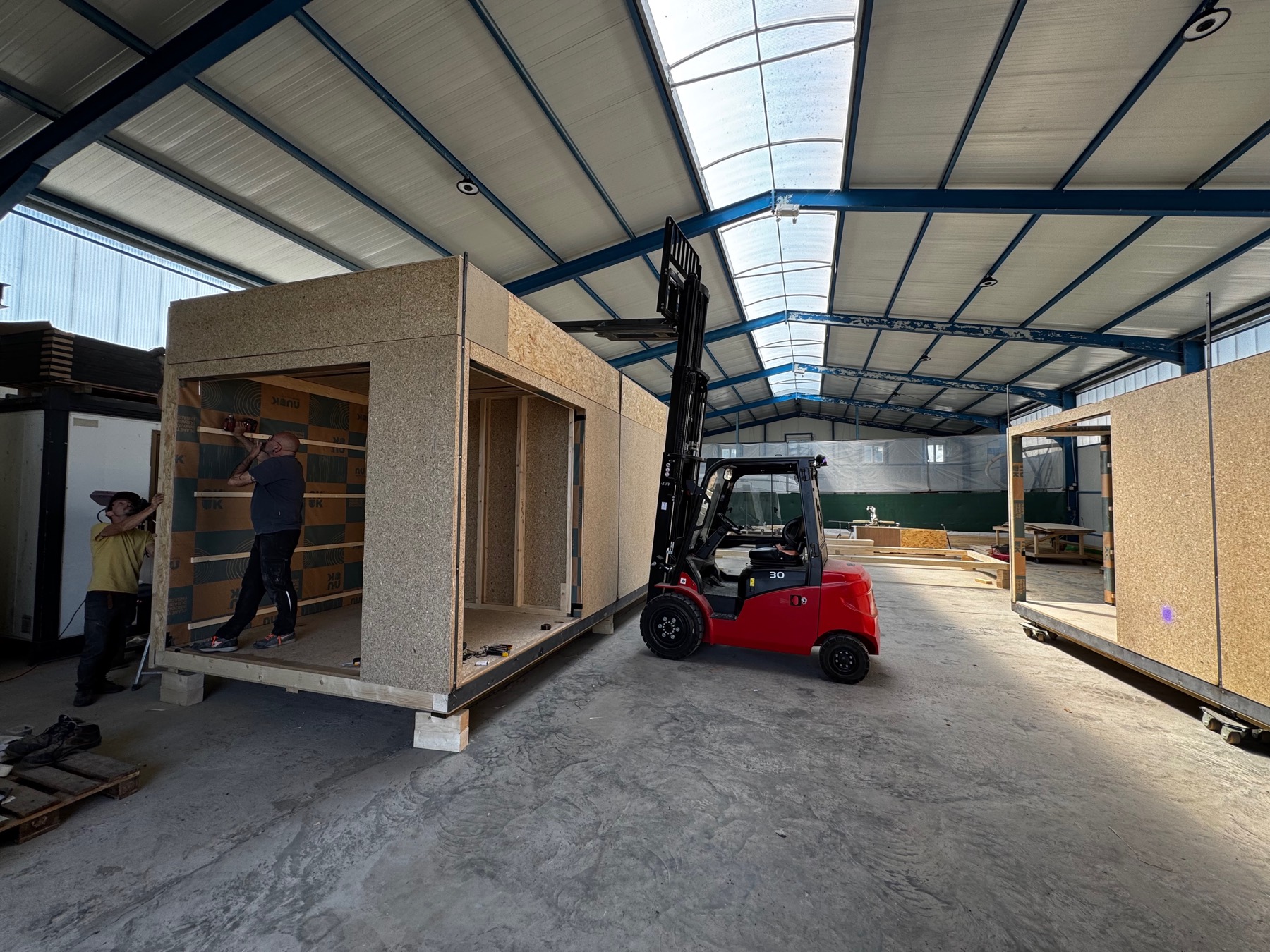 Deux compagnons manœuvrent au chariot élévateur un module en cours de montage, ossature bois et panneaux OSB apparents, sous la halle de production