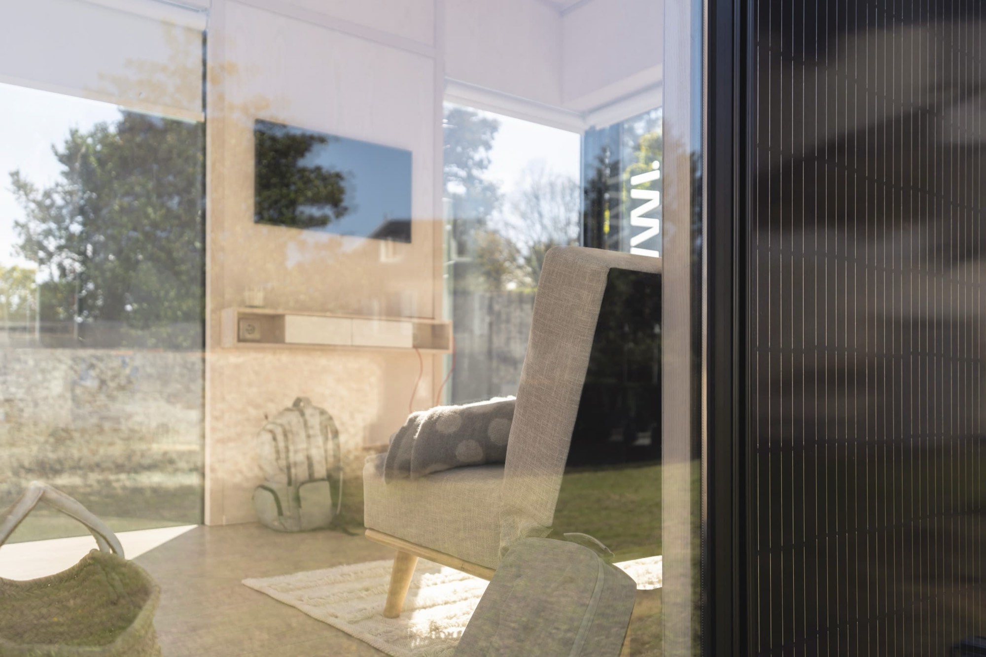 Séjour d'un module Hoummi vu à travers la baie vitrée : fauteuil et plaid, panier et sac à dos au sol, le verre superposant l'intérieur et le reflet du jardin