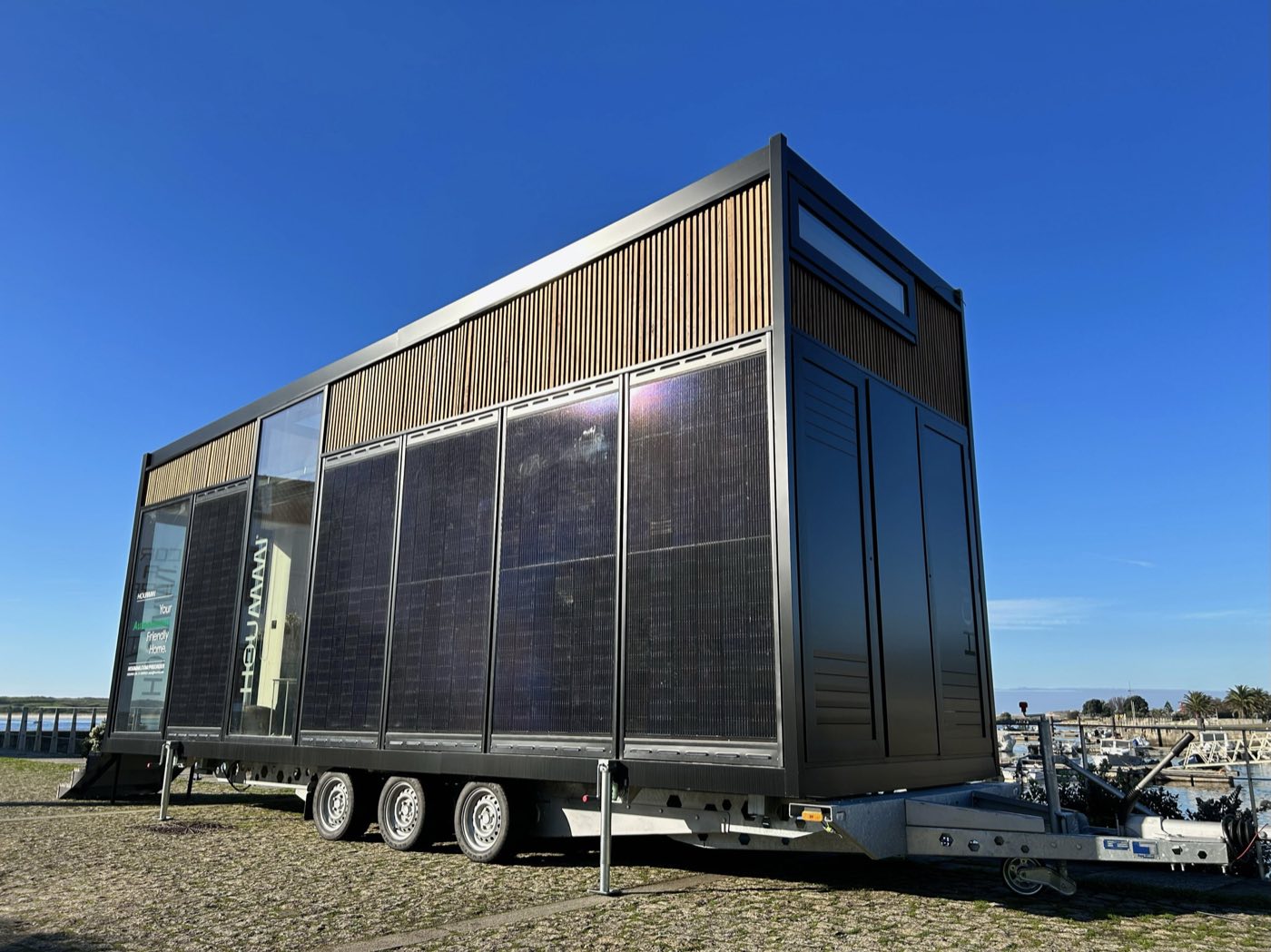 Tiny house Hoummi sur sa remorque à trois essieux, béquilles déployées, façade couverte de panneaux photovoltaïques et bardage bois en partie haute