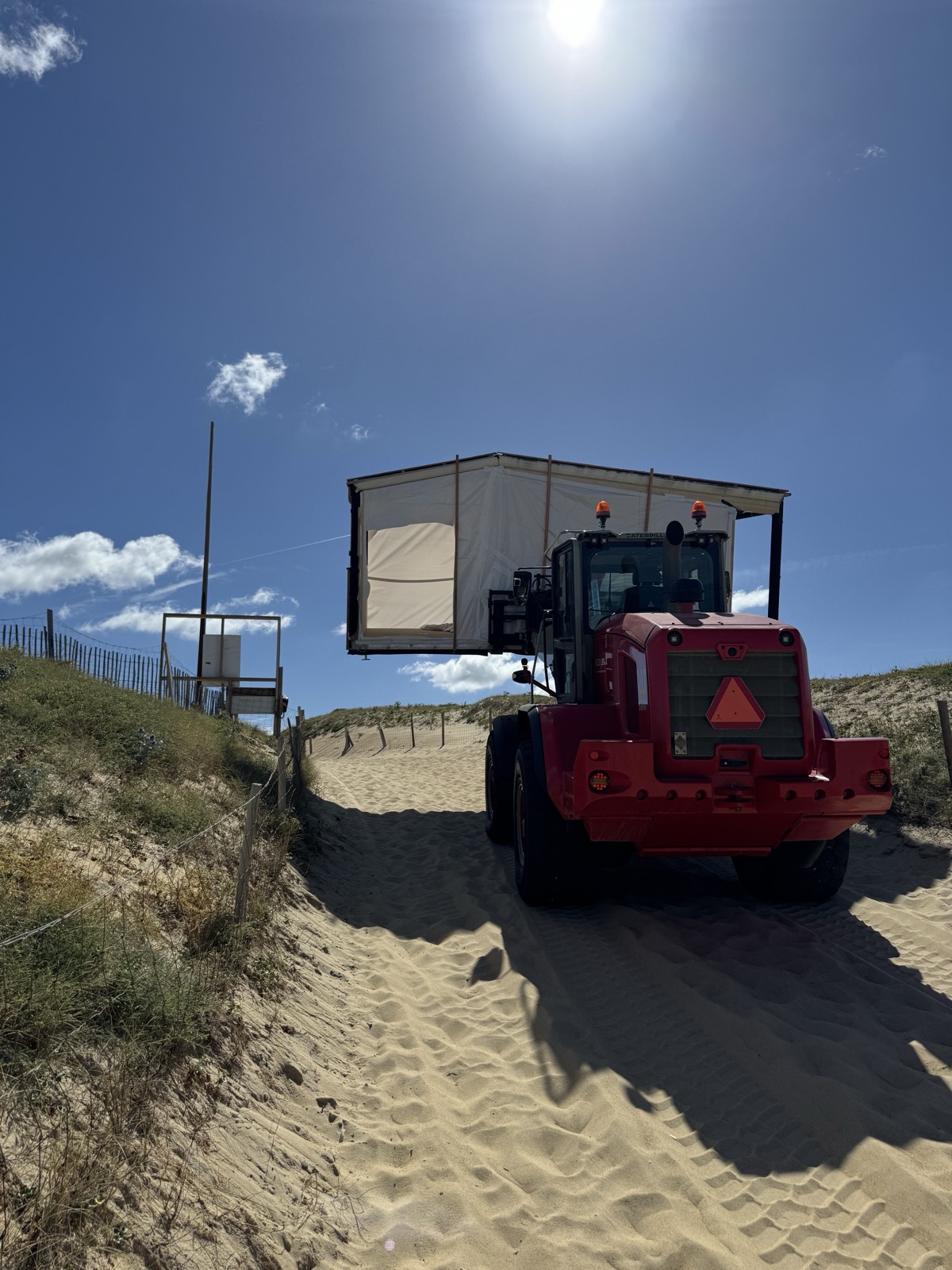 Une chargeuse télescopique rouge franchit la dune en portant un module encore sous emballage, ganivelles et végétation dunaire de part et d'autre du passage sableux