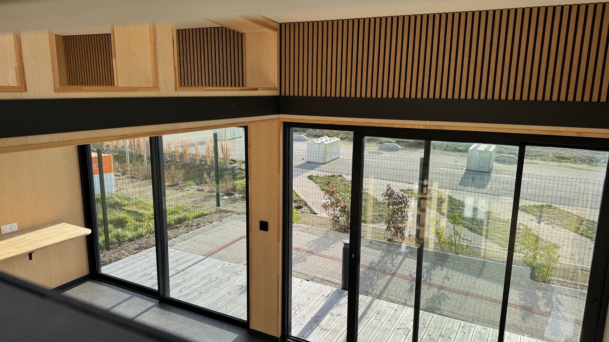 Intérieur d'un module Hoummi vu depuis la mezzanine : plan de travail mural en bois, paroi à tasseaux et baies coulissantes ouvrant sur une terrasse en bois